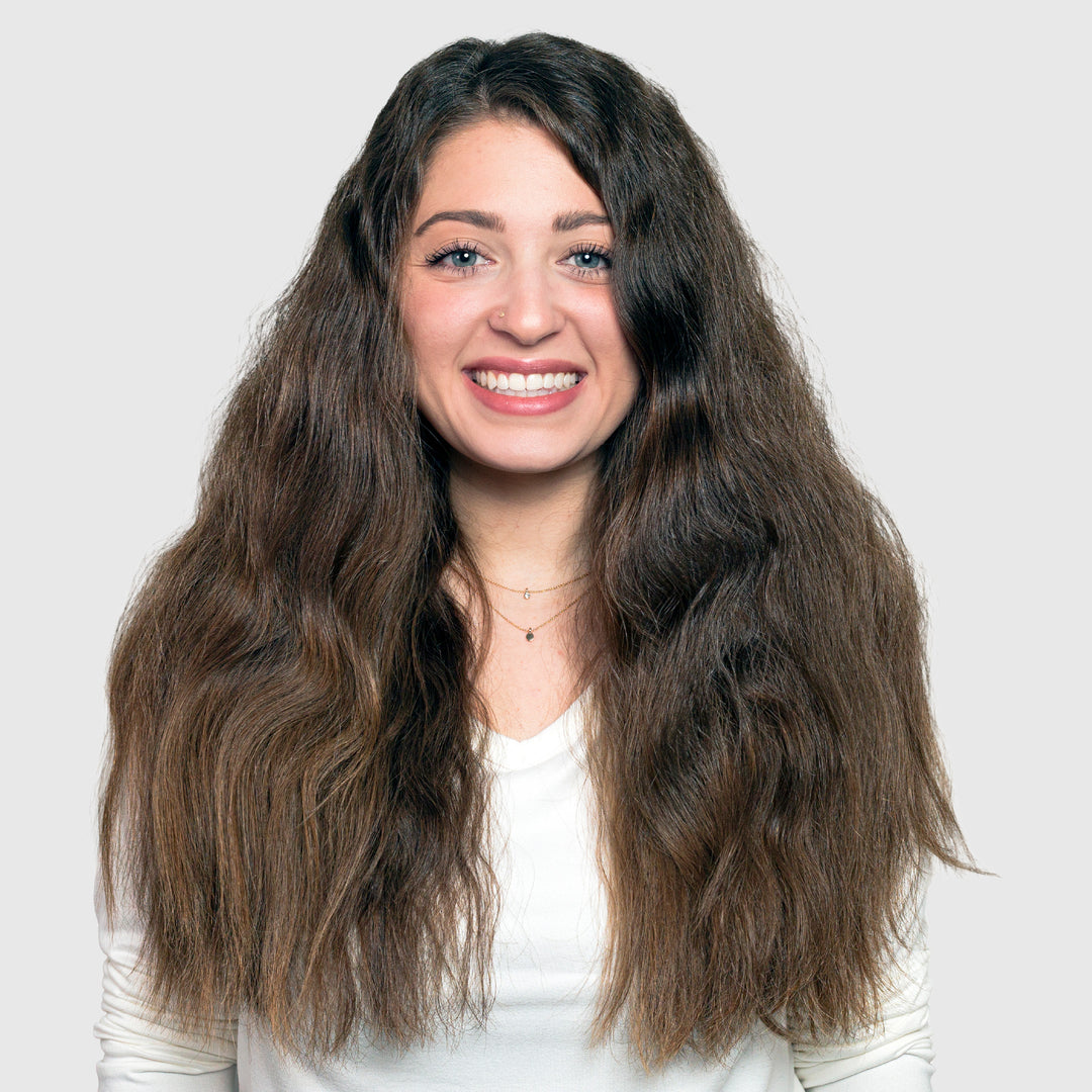 A woman with long, voluminous brown hair—radiant after using Calista Tools GLOSS Clear Shine Enhancement—and wearing a white top smiles at the camera against a plain light gray background.