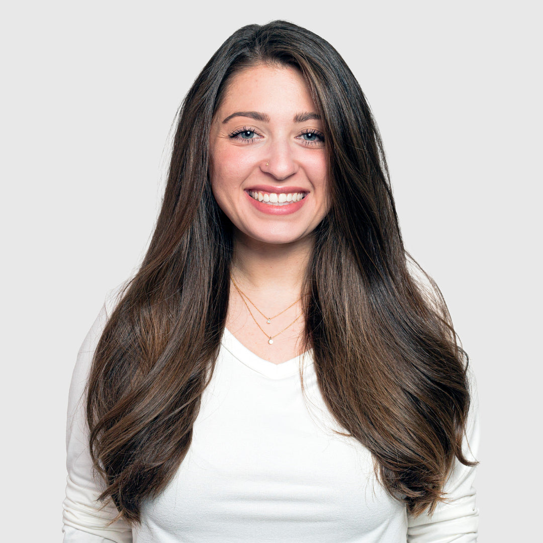 A young woman with long brown hair and a white top smiles at the camera against a plain gray background, her shiny locks showing the effects of Calista Tools GLOSS Clear Shine Enhancement.