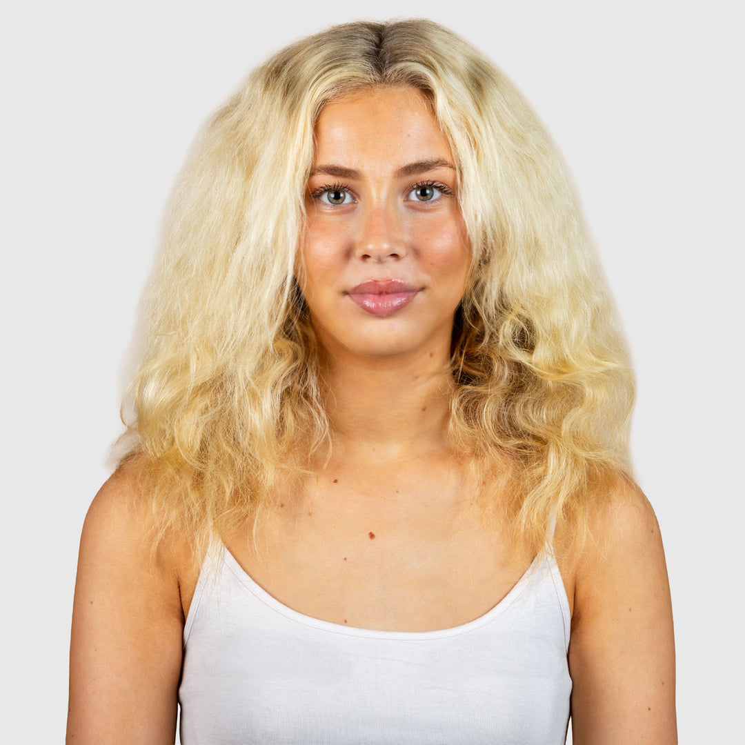 A young woman with long, wavy blonde hair and light skin, in a white tank top against a plain light gray background, looks directly at the camera with a neutral expression—her healthy locks cared for by JOUSSE Cleanse from Calista Tools.