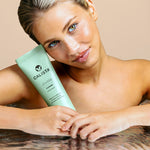 A woman with wet hair holds a green Calista Tools JOUSSE Condition tube, resting her arms on the water’s surface. She smiles softly, highlighting her healthy hair growth against a beige background.
