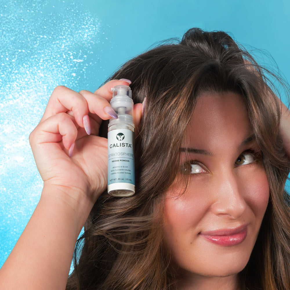 A woman with long brown hair holds a Calista Tools STRATOSPHERE Revive Powder near her head, smiling softly against a bright blue misty background, showcasing how to volumize hair with this talc-free formula.