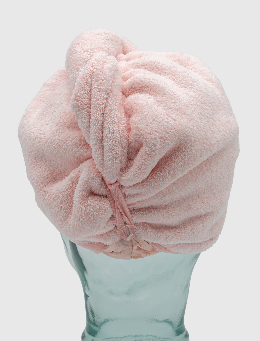 A glass mannequin head displays the Calista Tools Duet Satin-Microfiber Twist Towel in soft light pink, twisted and secured at the back. The background is plain and light-colored.