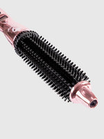A close-up of the metallic pink Perfecter Pro Long Hair Duo, a heated round brush with black bristles, photographed against a light grey background.
