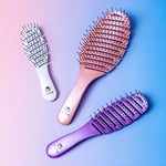 The Calista Tools SMOOTHIE Brush Trio includes three detangling brushes in white, pink, and purple with black-tipped bristles and branded handles—ideal for easy detangling and styling, showcased on a blue and pink gradient background.